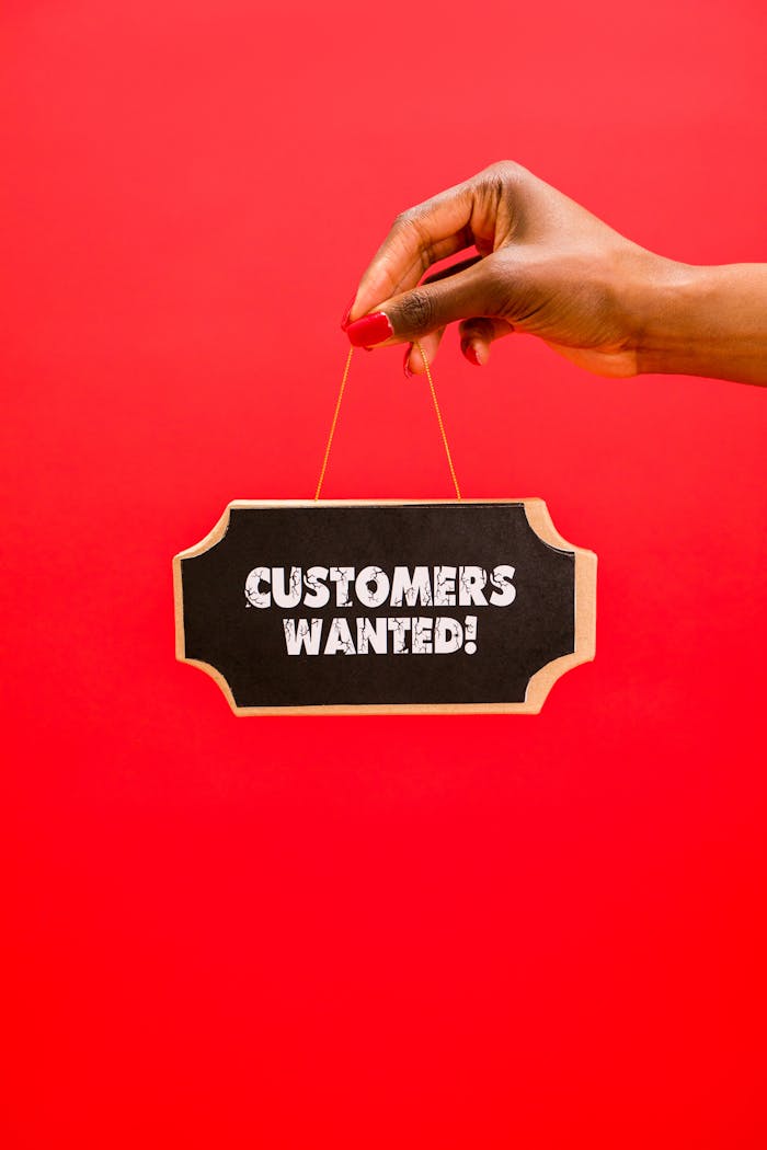 Close-up of a hand holding a 'Customers Wanted' sign against a bright red background.